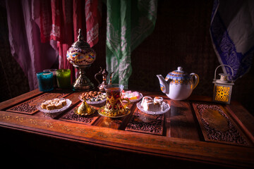 Arabian tea in glass with eastern snacks on vintage wooden surface. Eastern tea concept. Low light lounge interior with carpet. Empty space.