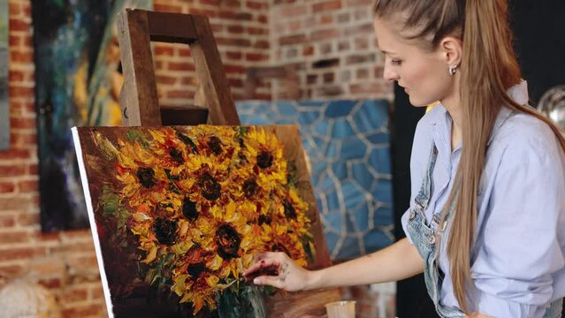 Young beautiful female artist painting still life with sunflower on canvas using oil paintings and palette knife. Painter creating artwork in art studio. Relaxation, leisure, hobby, stress management.