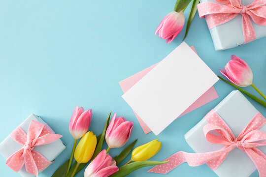 Mother's Day celebration concept. Flat lay photo of envelope with white card gift boxes and bouquet of pink tulips on isolated pastel blue background with empty space