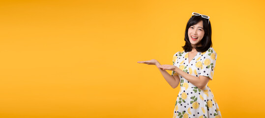 Portrait beautiful young asian woman wearing springtime dress and pointing to copy space for advertising isolated on yellow background. Summertime advertising banner concept.
