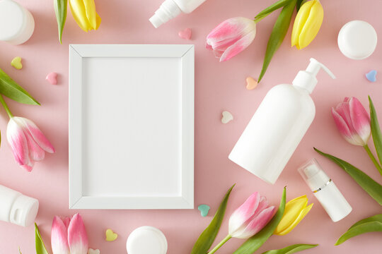 Natural cosmetic products concept. Top view photo of white photo frame pump bottle without label cosmetic tubes cream jars colorful hearts and yellow pink tulips on pastel pink background