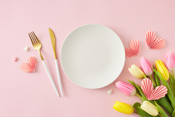 Mother's Day concept. Top view photo of white circle plate cutlery knife fork bouquet of flowers yellow pink tulips and paper hearts on isolated pastel pink background with blank space