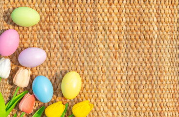 Happy Easter holiday greeting card concept. Colorful Easter Eggs and spring flowers on Papyrus rattan weave texture background. Flat lay, top view, copy space.