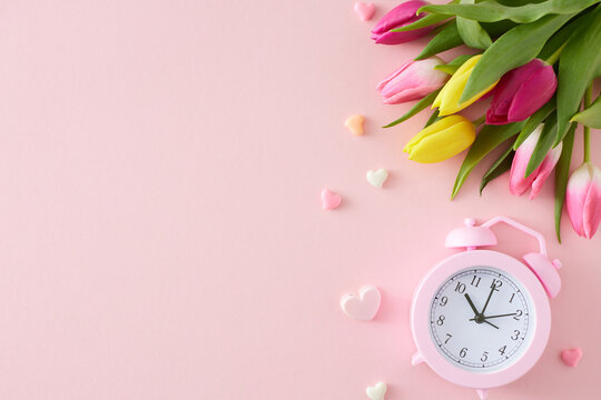 Mother's Day mood concept. Flat lay photo of bouquet yellow pink tulips alarm clock and colorful hearts on pastel pink background with empty space