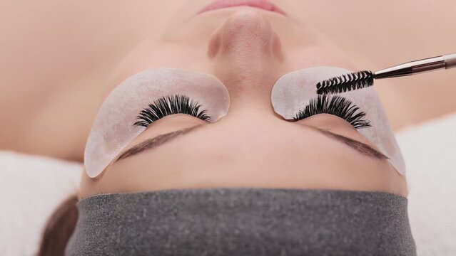 Eyelash extension procedure. Professional beauty master combing female lashes with special mascara brush. Fake eyelashes. Eyelashes extensions close up. Makeup artist and client in beauty salon.
