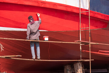 Asian painter on hanging platform is repainting hull of the old wooden fishing boat in shipyard...