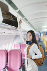 Young asian attractive woman travel by airplane, Passenger wearing headphone putting hand baggage in lockers above seats of plane