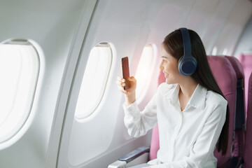 Young Asian attractive business woman passenger sitting on business class luxury plane while wearing wireless headphone and using smart phone mobile for relax during the flight