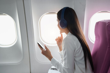 Young Asian attractive business woman passenger sitting on business class luxury plane while wearing wireless headphone and using smart phone mobile for relax during the flight