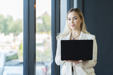 Young business woman standing and working in modern office, wearing suit and holding laptop computer in office area