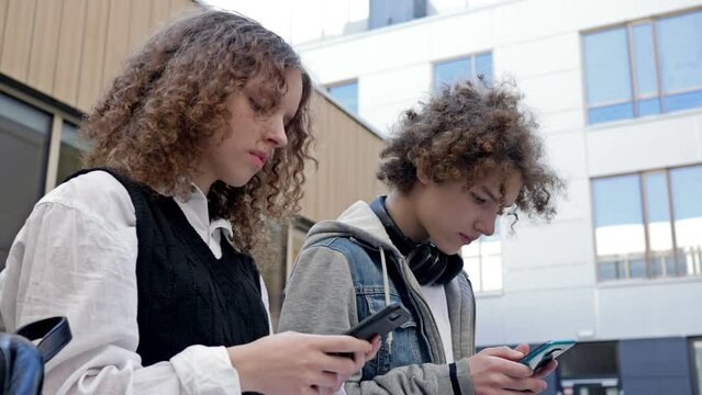 Two high school students, a boy and a girl, are sitting on a bench in the school yard. Each teenager has their own smartphone in their hands. Young people's passion for new technological trends.