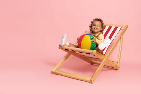 Happy African American Little Girl Smiling And Sitting On Beach Chair With Beach Ball Isolated On Pink Background, Two Years Old