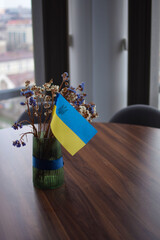 The Ukrainian flag stands in a vase of flowers on the table in front of the window.