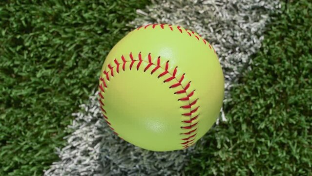 An overhead close-up shot of a softball sitting on the foul line of the outfield grass as the camera rotates – slow.