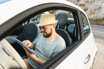 Naklejka premium Bearded man using digital tablet inside car while travel and road trip vacation holidays - road map and navigation concept