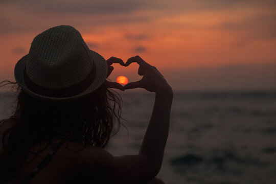 Traveler woman in hat shows image of shape heart with fingers at tropical sunset background, summer vacation. Lady tourist enjoy rest on seacoast. Travel relaxing holiday concept. Copy ad space