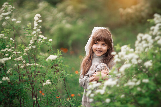 A cute little girl with brown hair holds favorite fluffy toy bunny among white flowers in the park. Background summer green flowering on the lawn. Child playing on the spring nature outdoors. Soft