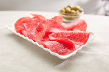 salami slices on a white plate with olives in the background. sausage close-up. meat delicacy on a sunny day. traditional mediterranean snack on a light surface. dried sausage serving. antipasta macro