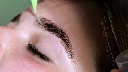 Beautician brushes painted eyebrows of female client doing lamination. Master uses brush for long-lasting styling in beauty salon closeup