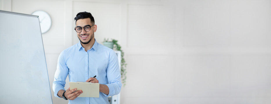 Cheerful confident arab millennial guy teacher in glasses stands near whiteboard in office class interior, panorama