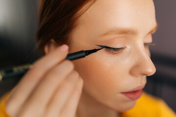 Closeup face of red-haired young woman using contour brush drawing arrows on eyelids looking at...