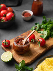 Sauce paste harissa with pepper and tomato in a jar on a dark background with fresh vegetables and herb. Adjika. Traditional italian, Georgian and Arabic cuisine. Homemade food..