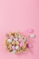 Easter candy chocolate eggs and almond sweets lying in a bird's nest decorated with flowers and feathers on pink background. Happy Easter concept.