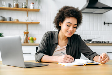 Positive african american girl, with short curly hair, student or freelancer, sitting in the kitchen at a table with a laptop, studying or working online, takes notes in a notebook, smiles