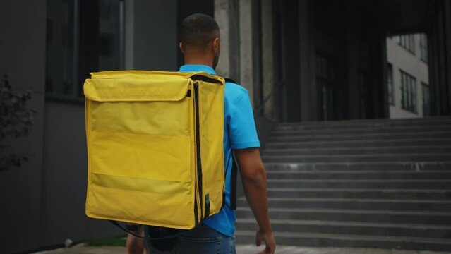 Back View of the African American Courier Finding the Customer Location. Multiethnic Man Delivery Going To the Buyer Workplace Carrying the Yellow Backpack Wearing Blue T-Shirt