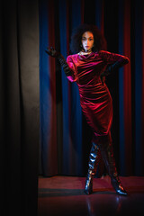 Full length of stylish african american woman in dress and knee boots posing on red background with blue light.