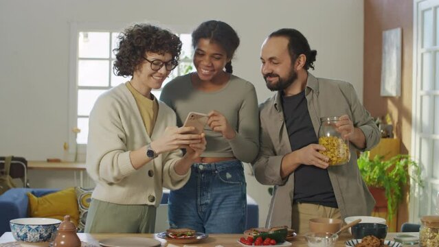 Full medium shot of Caucasian young woman, black young girl and Caucasian man standing in front of table. All three smiling for selfie, girl putting cherries on eyes, then looking at photo, laughing