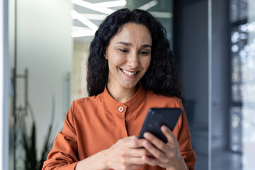 Beautiful and successful Hispanic woman at work smiling and looking at the phone screen, businesswoman in the office holds smartphone in her hands closeup browses the Internet uses application call