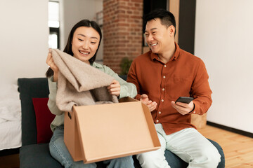 Excited asian couple unpacking cardboard box at home, man and woman buyers opening cardboard parcel...