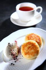 Cottage cheese pancakes with sour cream on a white plate. Dark background. Sweet breakfast cheese pancakes. A cup of tea.Vertical photo.