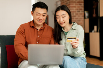 Smiling korean middle aged man and young woman using credit card and laptop, sitting on sofa in living room interior