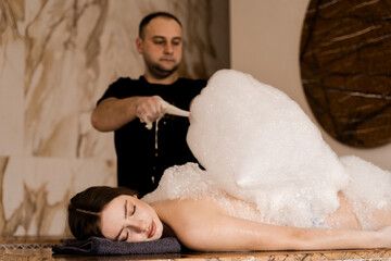 Attractive girl relaxing in spa. Masseur is making foam peeling in Turkish spa hammam.