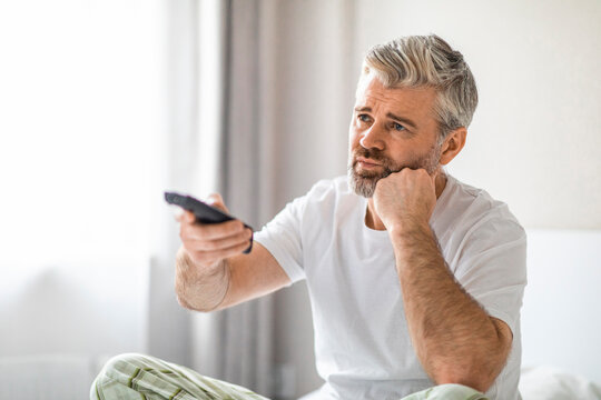 Bored Mature Man Sitting On Bed At Home, Watching Tv