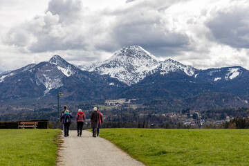 A group of elderly people walking along a village road, between green fields towards the Alps