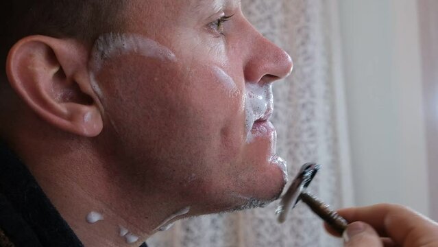 A man shaves with an old style razor with a replaceable blade and a foam shaving brush. Traumatic shave. The old way of skin and beard care. Bristle haircut. Soaped face close-up