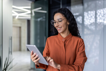 Latin american female programmer inside office standing near window using tablet computer for...