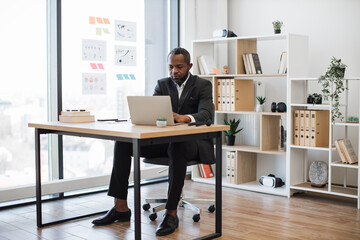 Thoughtful businessman in formal clothes typing on portable computer while sitting at office desk...