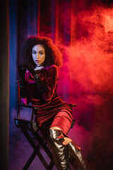 Stylish african american model in dress sitting on chair on red background with blue light and smoke.