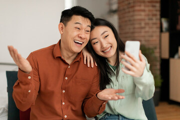 Excited asian couple taking selfie on smartphone, communicating via video call, talking and smiling at webcamera