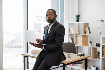 Experienced african american male employer working on modern laptop computer while relaxing on edge of office desk. Focused businessman in formal wear checking emails using online application.