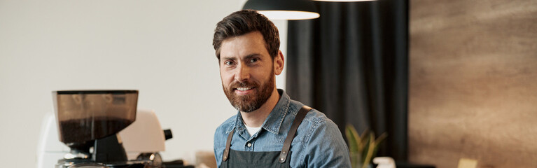 Handsome Barista with stylish beard wearing apron smiling while behind a counter in the coffeeshop