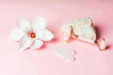 Obraz na płótnie Canvas Rose quartz crystal facial roller and gua sha scraper, magnolia flower on pink background. Facial massage kit for lifting therapy . Skin care anti-aging tools