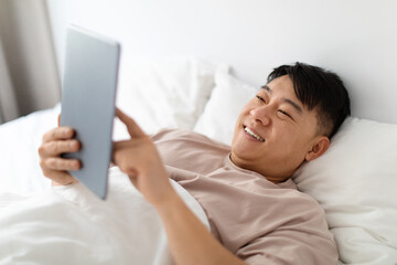 Positive korean man using modern digital tablet in bed