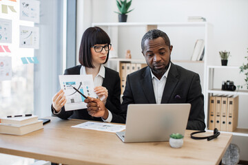 Businesspeople in formal wear looking into computer webcam while working with data visualizations in creative workplace. African american male and caucasian female brainstorming online in office.