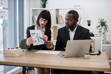 Thoughtful multiethnic businessman and his caucasian colleague arranging video conference using bar graphs held by woman. Competent professionals holding remote team meeting via laptop application.