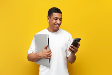 young african american guy in white t-shirt holding laptop and using smartphone on yellow isolated background